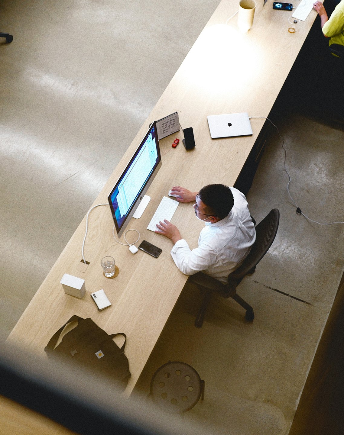 A man wearing glasses works at a computer on a wooden desk.